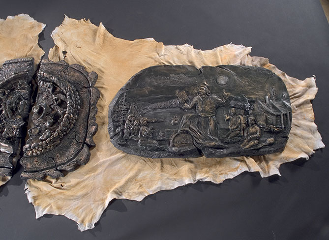 The right side of the work shows a priest from the Catholic Church preaching to his faithful who are kneeling with their hands folded in prayer. He holds his bible (the book) high as if he were imposing a new law, as he crushes a teueikan, the sacred drum of the Innu, underfoot. The patinated plaster imitates bronze: greenish tones embellished with golden highlights.